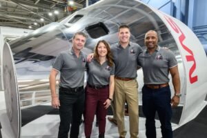 The four Artemis II astronauts pose for a photo in front of an Orion simulator. The crew members are in matching grey polo shirts with their mission insignia.