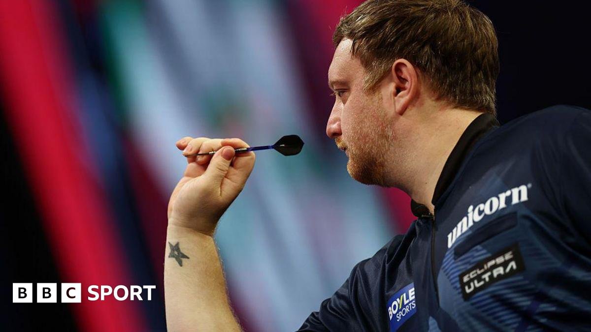 Close-up photograph of Cameron Menzies as he is about to throw a dart with his right hand. He has short brown hair and is wearing a navy blue top.