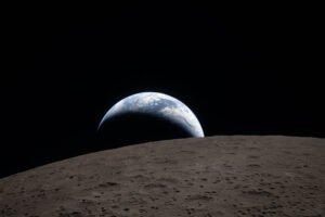 A muted blue Earth with bright white clouds sets behind the cratered lunar surface. The dark portion of Earth is experiencing nighttime. On Earth’s day side, swirling clouds are visible over the Australia and Oceania region. In the foreground, Ohm crater has terraced edges and a flat floor interrupted by central peaks. Central peaks form in complex craters when the lunar surface, liquefied on impact, splashes upwards during the crater’s formation.
