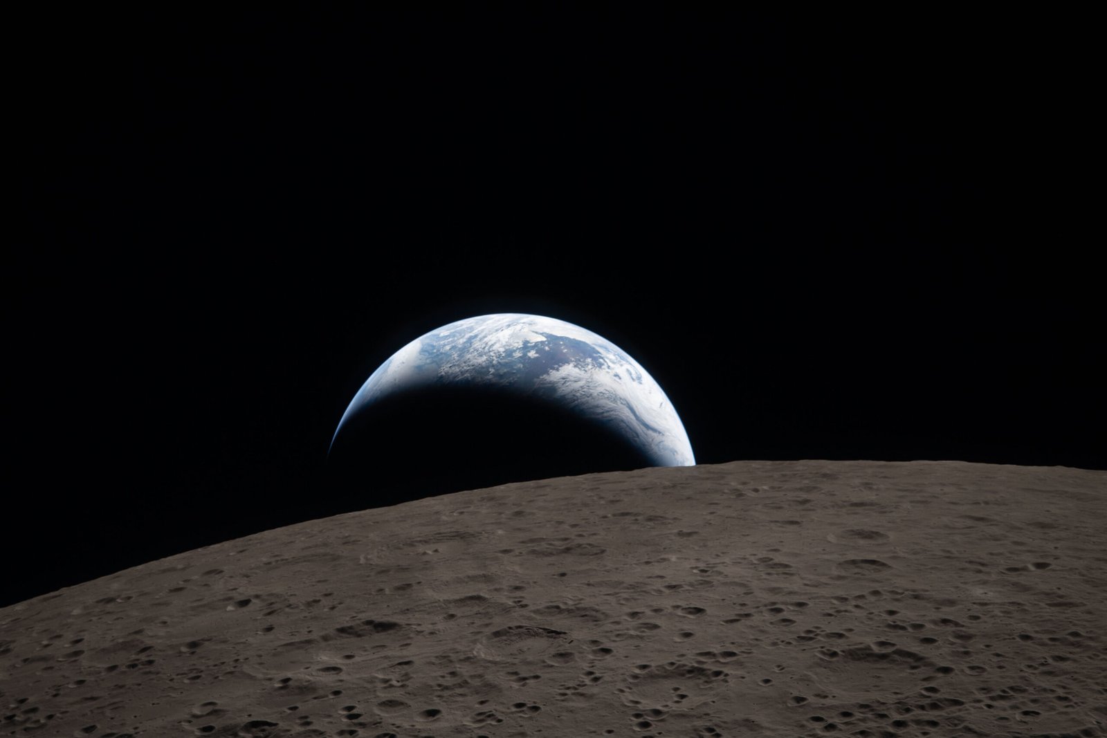 A muted blue Earth with bright white clouds sets behind the cratered lunar surface. The dark portion of Earth is experiencing nighttime. On Earth’s day side, swirling clouds are visible over the Australia and Oceania region. In the foreground, Ohm crater has terraced edges and a flat floor interrupted by central peaks. Central peaks form in complex craters when the lunar surface, liquefied on impact, splashes upwards during the crater’s formation.