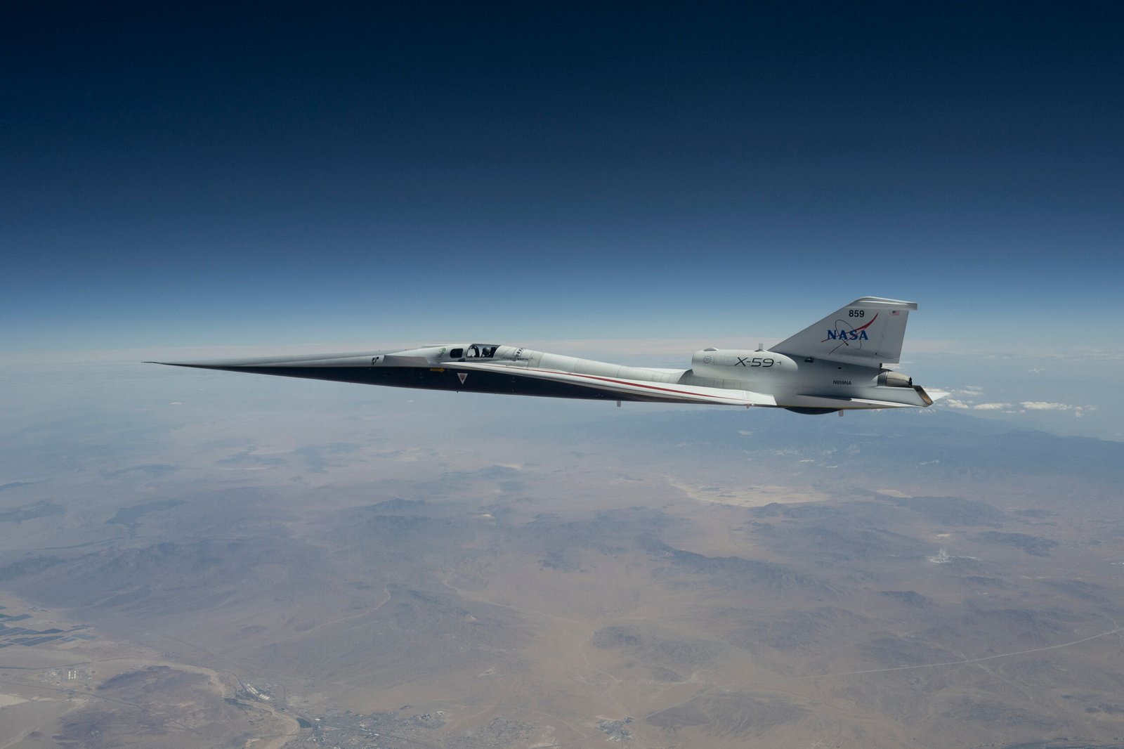 NASA’s X-59 quiet supersonic research aircraft flies above the Mojave Desert on a clear day. The aircraft is white with light gray, red, and blue accent colors. A NASA logo is visible on its tail, along with the number 859 above it.