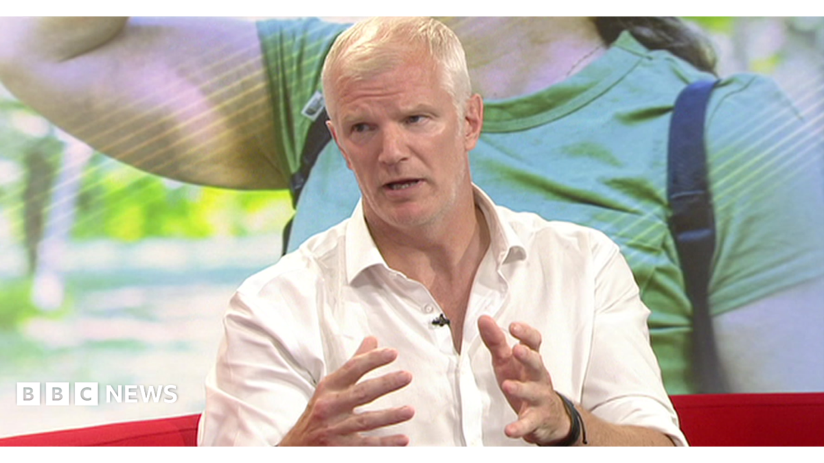 A man with short white hair is wearing an open-neck white shirt and sitting on a red sofa in a TV studio. There is a photographic backdrop behind him illustrating the heatwave, with a woman in summer clothes and a backpack.