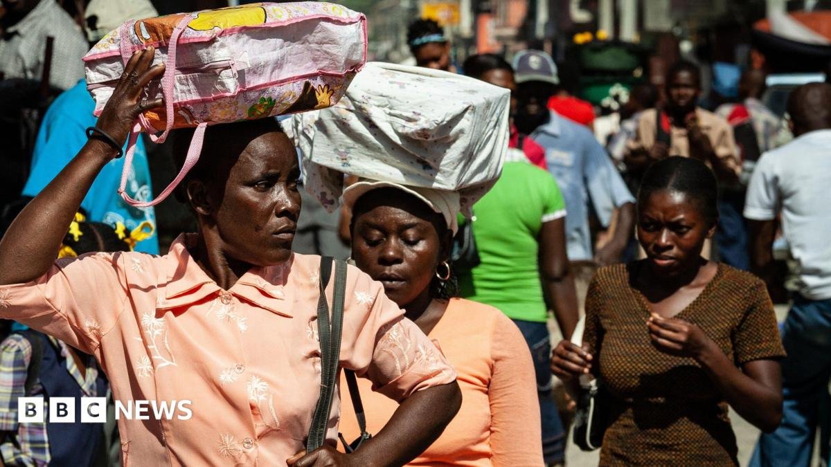 Three Haitian women carry bags on their heads as they walk down a crowded street. Two women wear orange blouses and one wears a brown t-shirt.