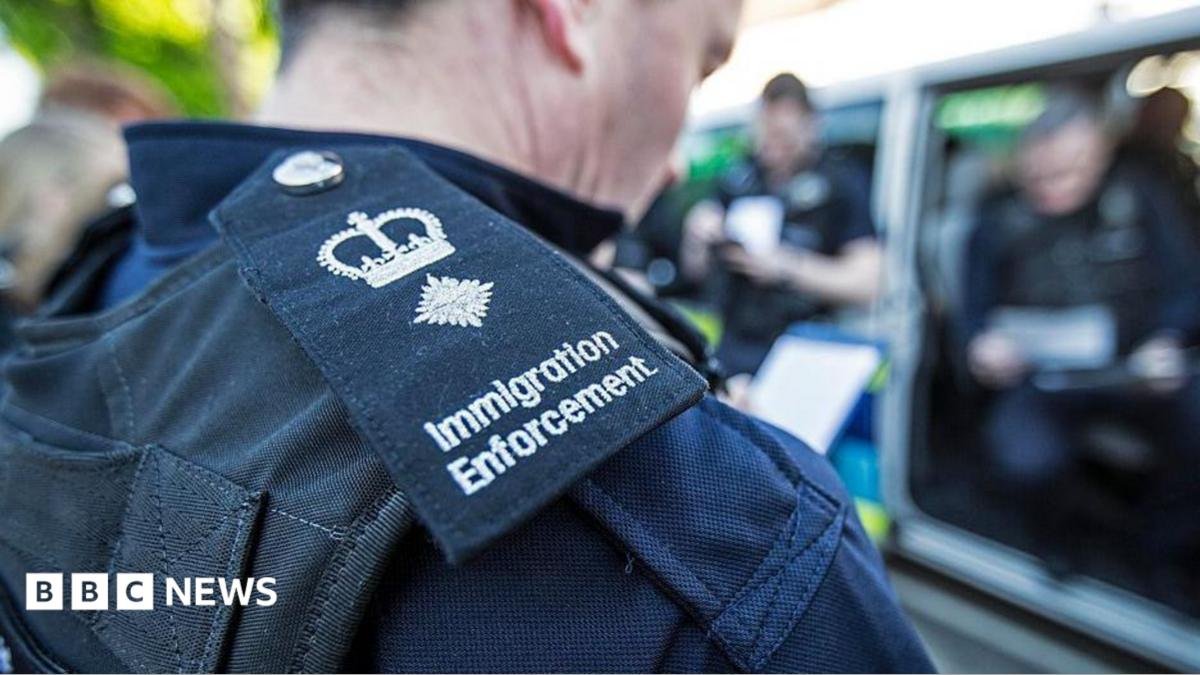 A close up of the lapel badge of an immigration enforcement officer. Other officers are blurred in the background.