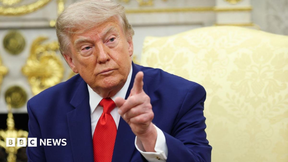 President Donald Trump pointing his finger. He has a serious look on his face and is wearing a blue suit with a white shirt and red tie.