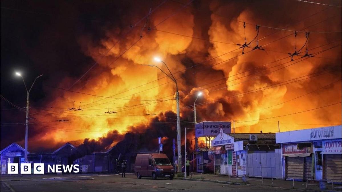 Large fire engulfs a market area at night, sending towering flames and thick smoke into the sky above nearby buildings, streets, and overhead power lines.