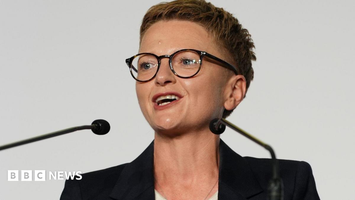 Bev Craig has short spiky red hair, dark glasses and wears a dark jacket with white roll-neck top.