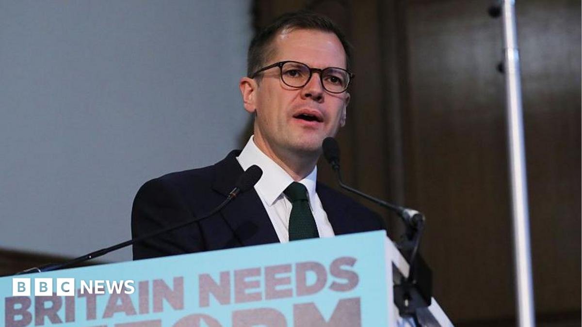 Headshot of Jenrick who speaks into two small microphones on stands on a podium in front of him that has a blue sign partially cut out of the frame, but you can read it says 'Britain needs reform'.