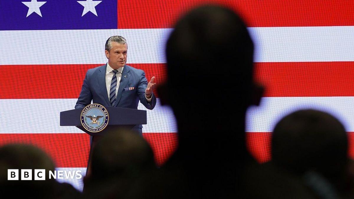 Pete Hegseth stands in front of a podium with an American flag projected behind him. There are silhouettes of a crowd watching him in the foreground. He is gesturing and smiling, wearing a blue suit and striped tie.