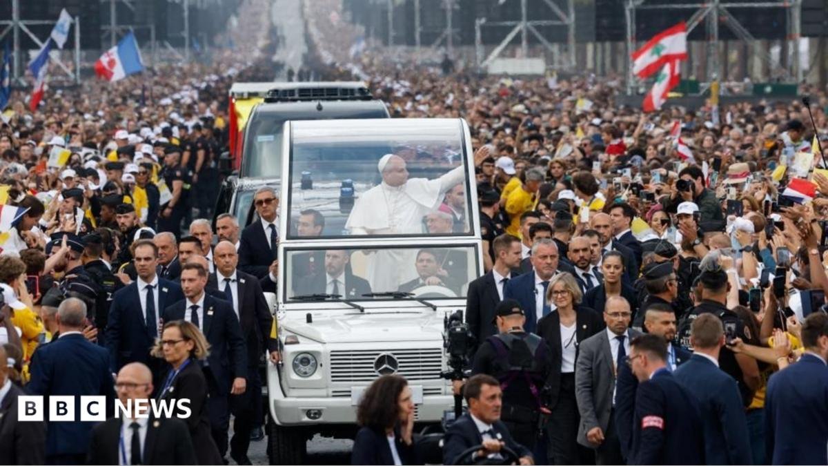 Pope Leo smiles and holds his arm up in his popemobile. Crowds can be seen waving flags and gesturing towards the pope. Some are holding up their phones or cameras to take pictures