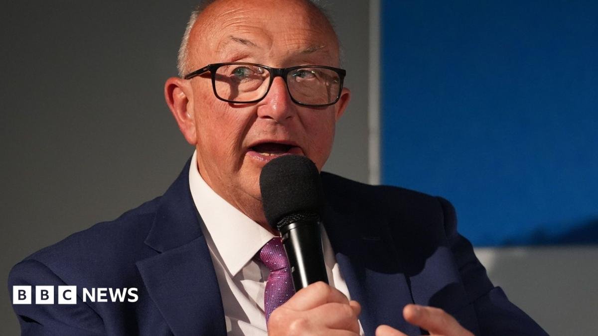 Colin Sutton is wearing black rimmed glasses and a navy suit, white shirt and purple patterned tie. He is holding a microphone close to his mouth and looks to be in a corporate environment.